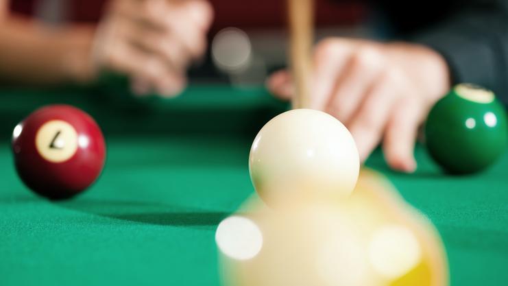Nahaufnahme einer weißen Billardkugel und einer roten Kugel auf einem grünen Billardtisch, Hände im Hintergrund.