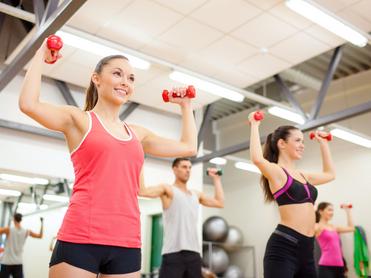 Vier Personen heben Gewichte in einem Fitnessstudio. Zwei Frauen tragen Sporttops, ein Mann hat ein T-Shirt an.