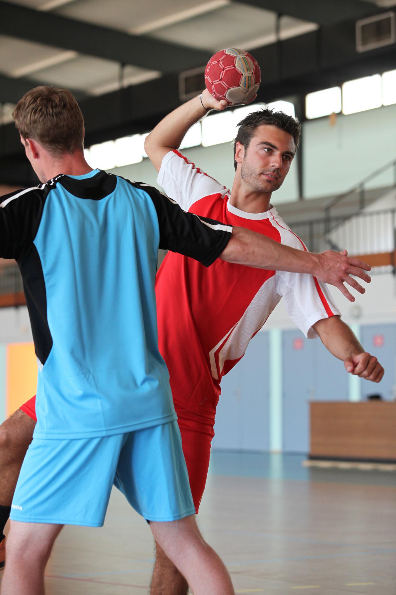 Zwei Handballspieler in action, einer wirft den Ball, während der andere ihn blockt. Halle mit Holzboden im Hintergrund.